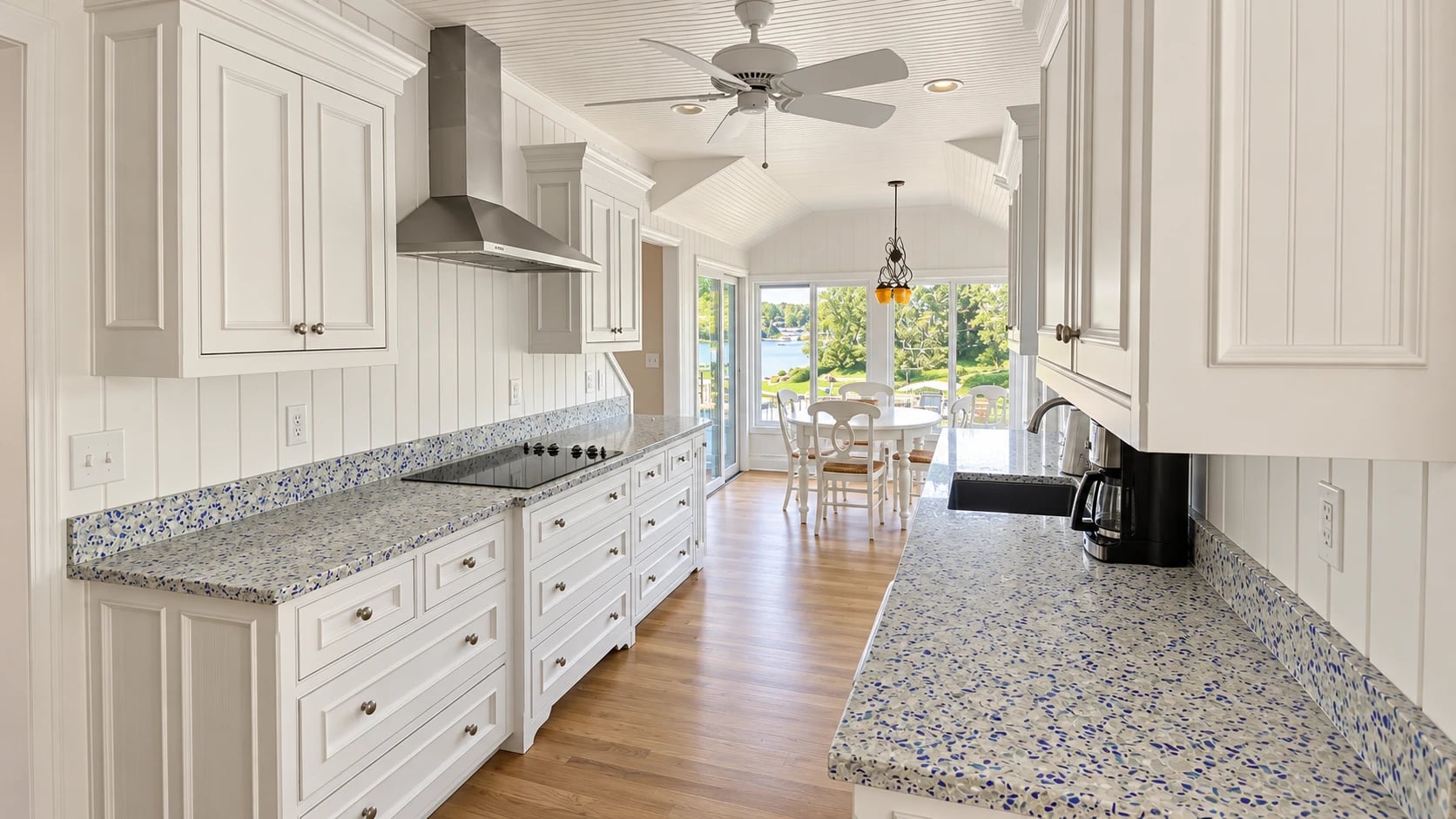 White galley kitchen with blue-speckled countertops, recessed ceiling lights and a dining area beside large windows